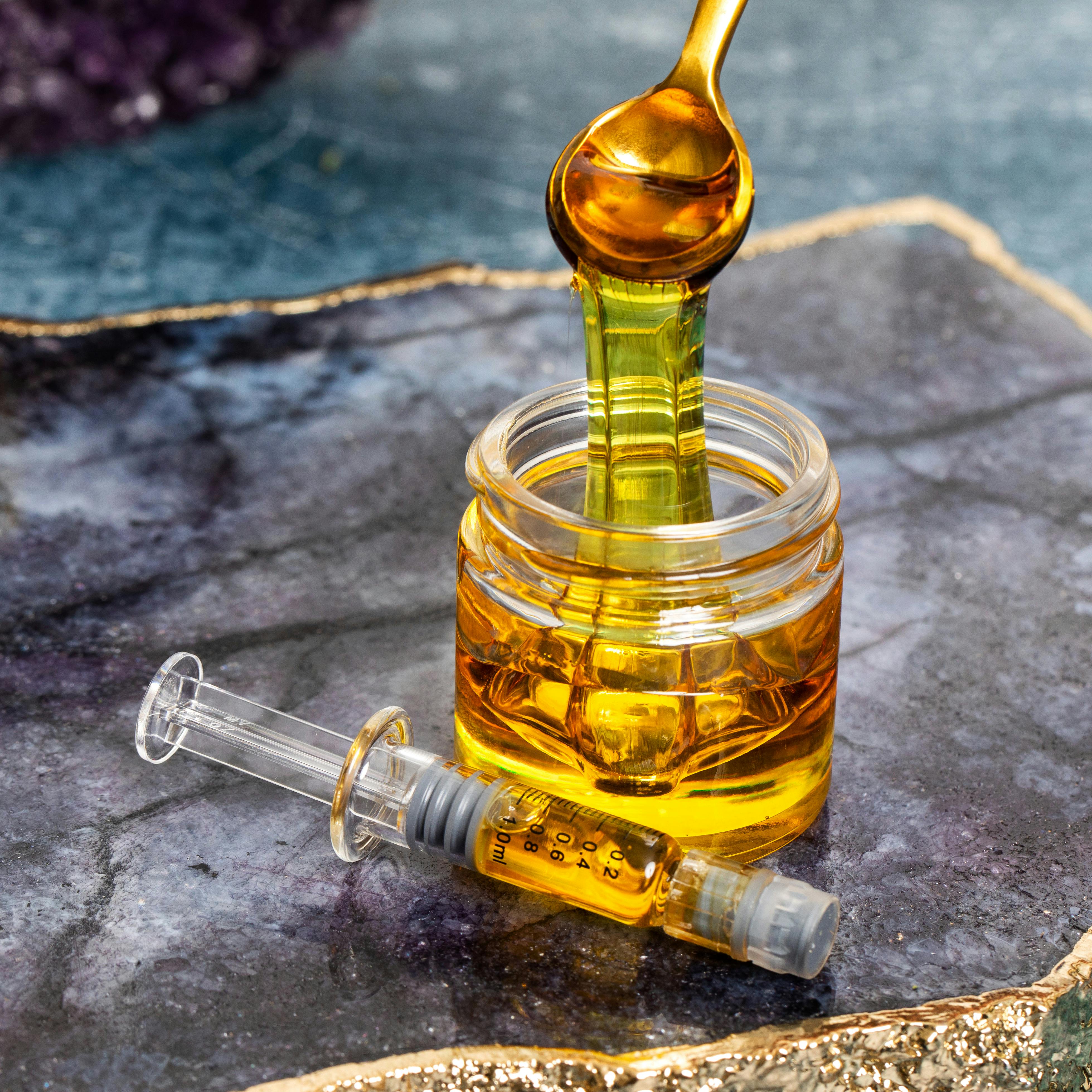 A thick stream of golden distillate drips from a spoon into a small glass jar filled with viscous amber oil, with a clear dosing syringe containing the same liquid resting beside it on a textured stone surface.”