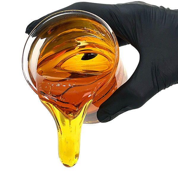 A hand wearing a black glove pours thick, golden amber distillate from a glass beaker, with the viscous liquid flowing downward in a smooth stream.